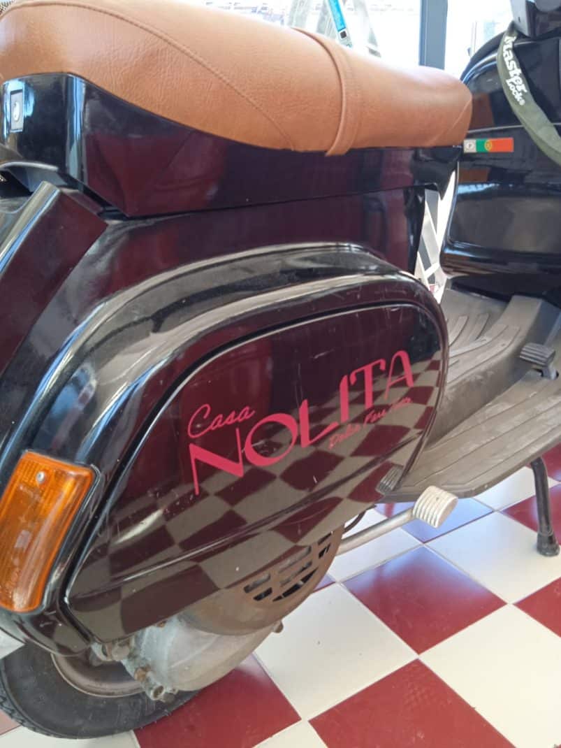 Logotipo Casa NOLITA na lateral de uma scooter Vespa preta com assento marrom.