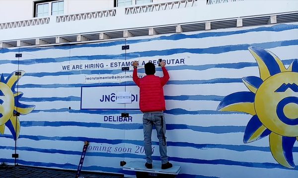 Vinil aplicado em tapumes com design de ondas azuis e sol sorridente, promovendo ambiente vibrante e.