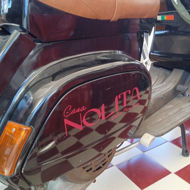 Logotipo Casa NOLITA na lateral de uma scooter Vespa preta com assento marrom.