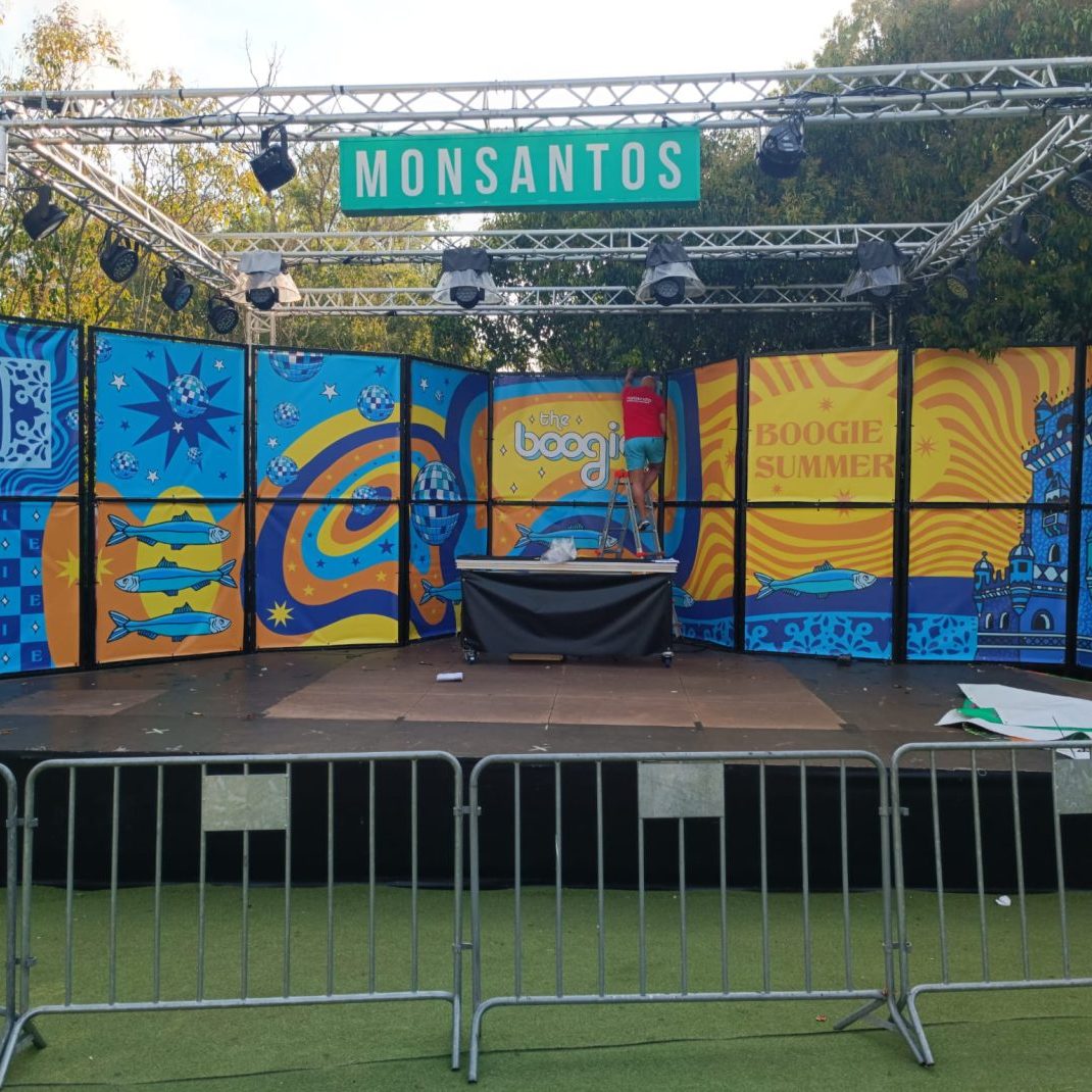 Palco de evento ao ar livre com painel colorido e sinalização "Monsantos" no topo, preparado para sh.